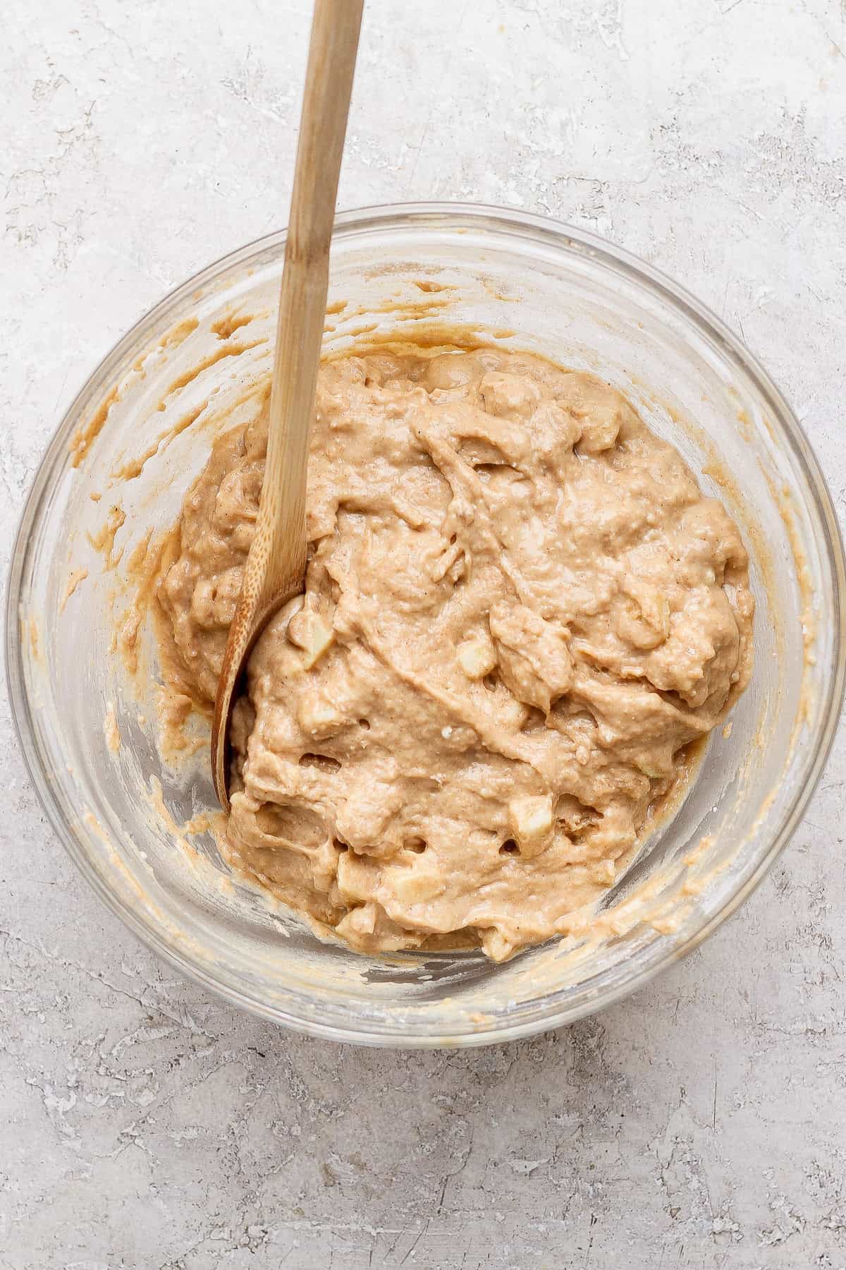 A glass bowl containing thick, chunky batter and a wooden spoon, set on a light-textured surface.