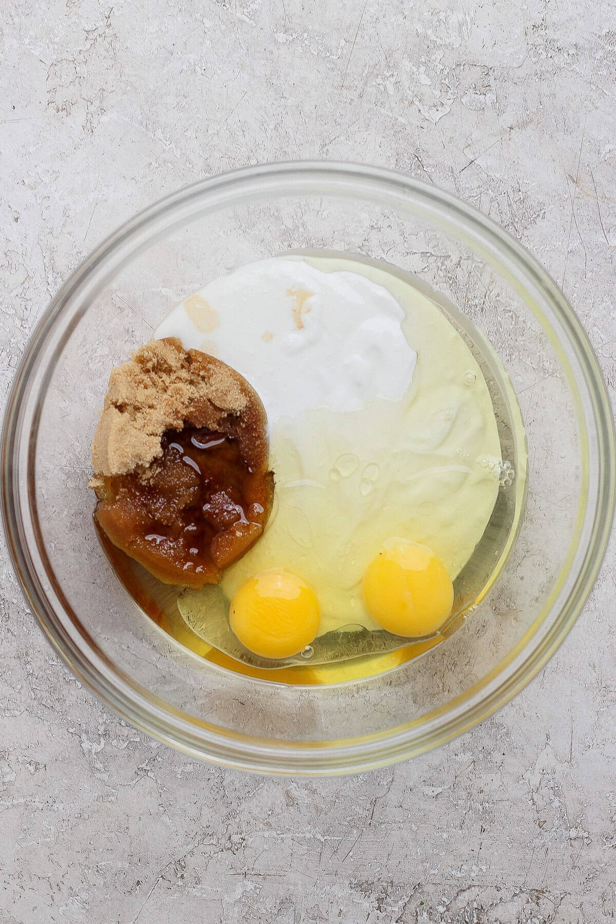 A glass bowl containing two eggs, brown sugar, honey, and a white liquid ingredient on a light textured surface.