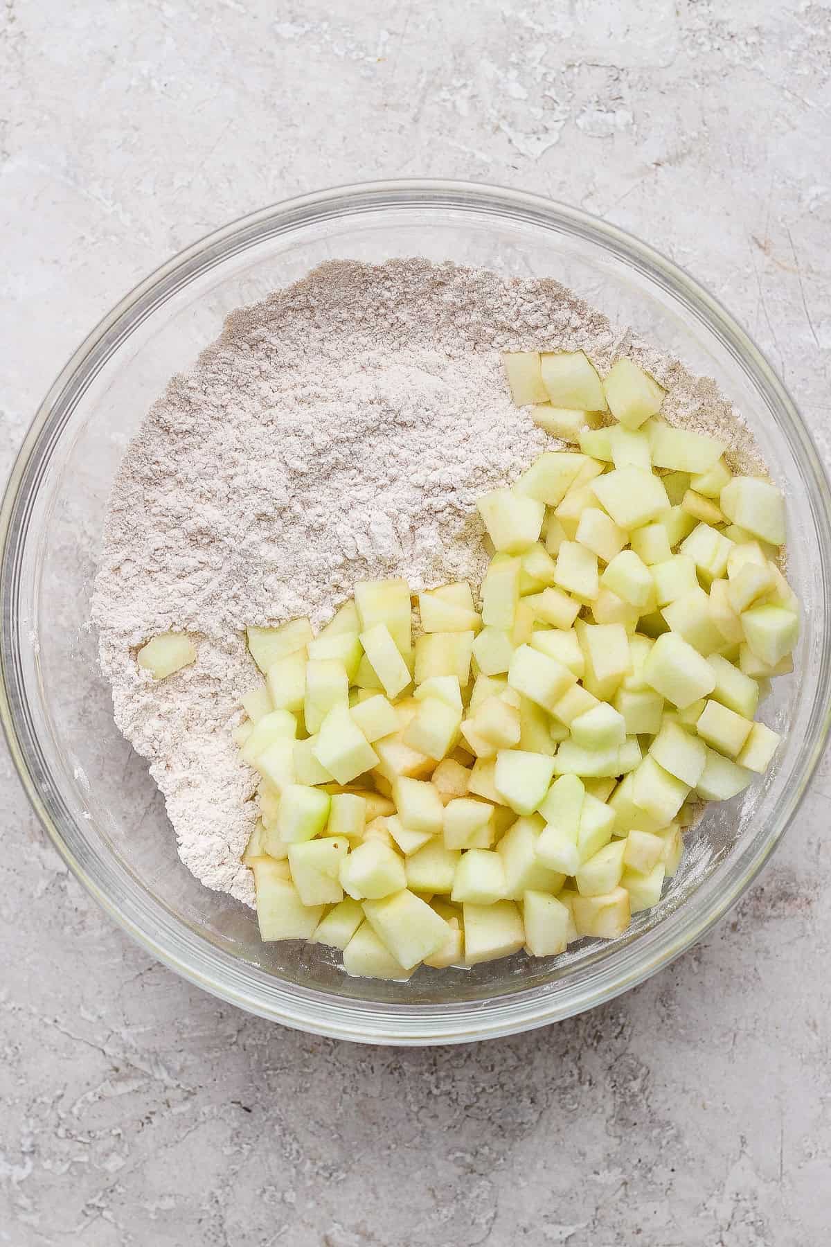 A glass bowl containing flour and diced green apples on a light textured surface.