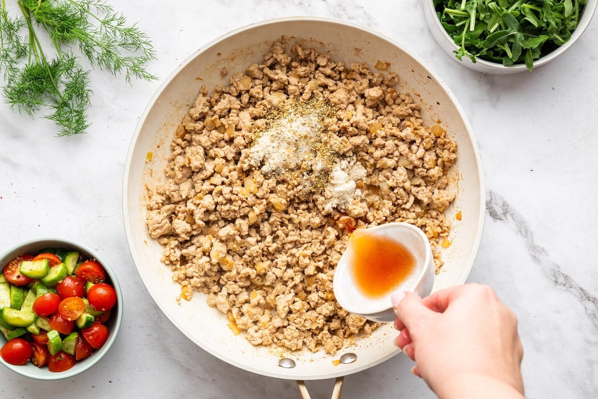 A womans hand pouring red wine vinegar into a skillet with ground turkey, herbs, and spices.
